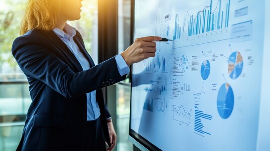 A woman in a suit pointing at a large monitor displaying graphs