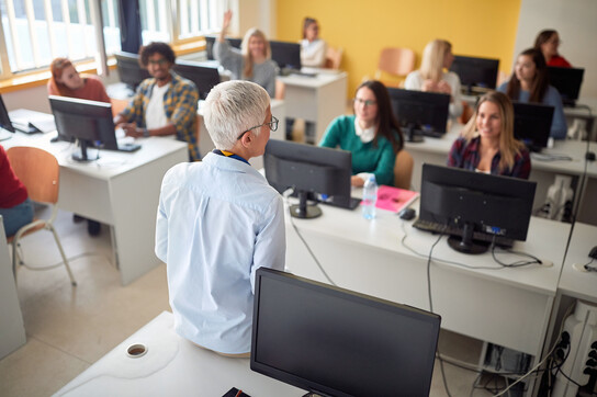 Eine Dozentin steht vor der Klasse mit jungen Menschen an PCs